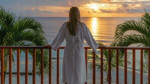 A person standing on a balcony overlooking a sunset over the ocean, with palm trees and wooden furniture visible. - Hopkins Bay Belize, a Muy'Ono Resort (Hopkins)