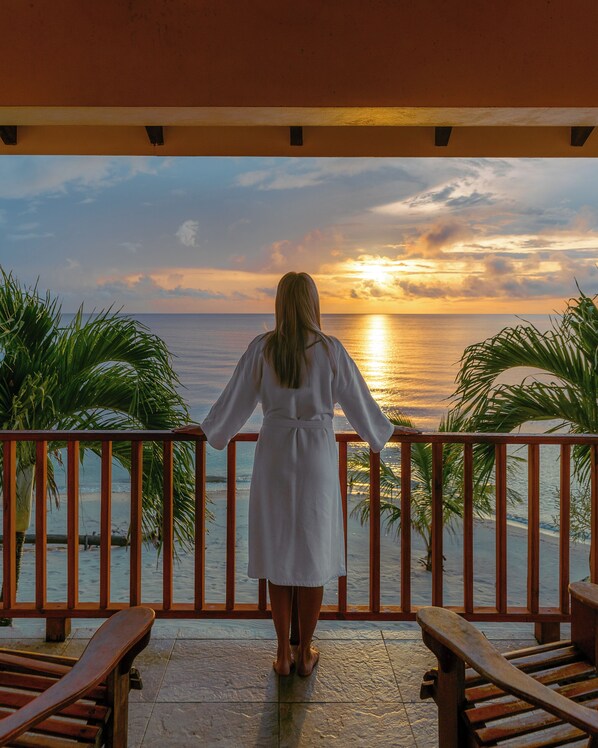 A person standing on a balcony overlooking a sunset over the ocean, with palm trees and wooden furniture visible.