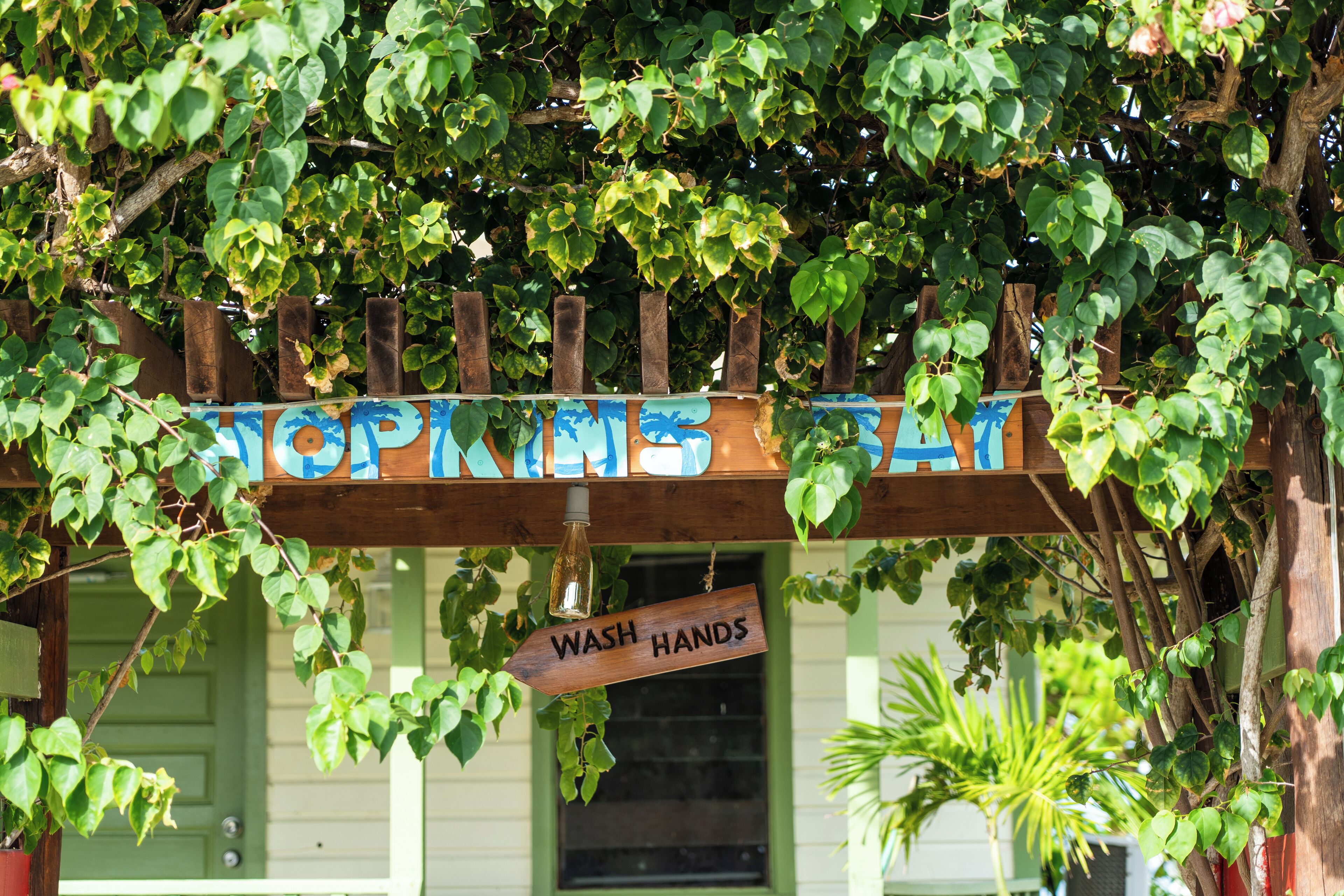 Een houten bordje met daarop 'WASH HANDS', hangend onder een houten pergola met groene bladeren.