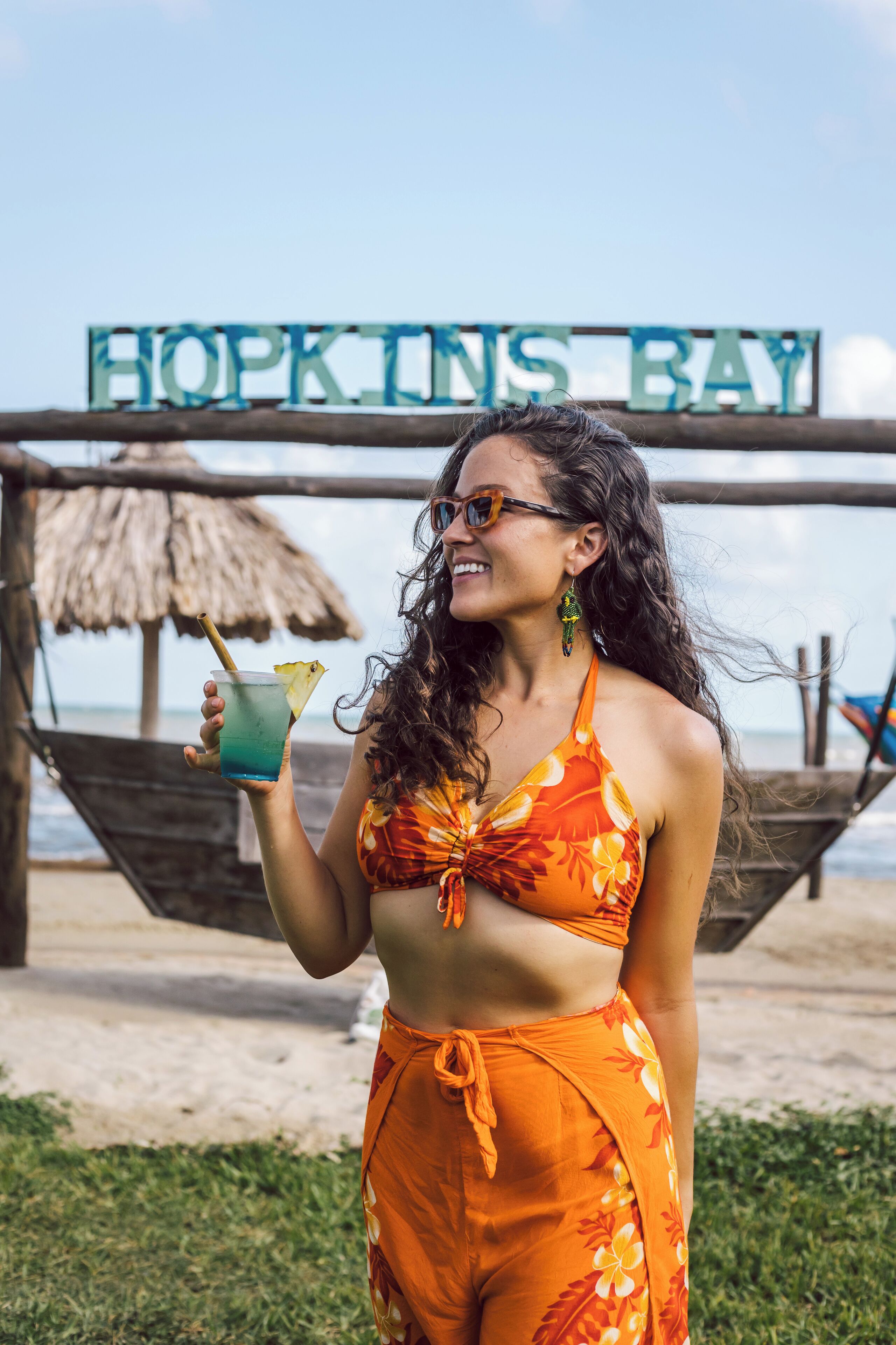 A person holding a drink with a straw and a slice of lime, wearing a floral bikini, standing in front of a sign that reads "HOPKINS BAY".
