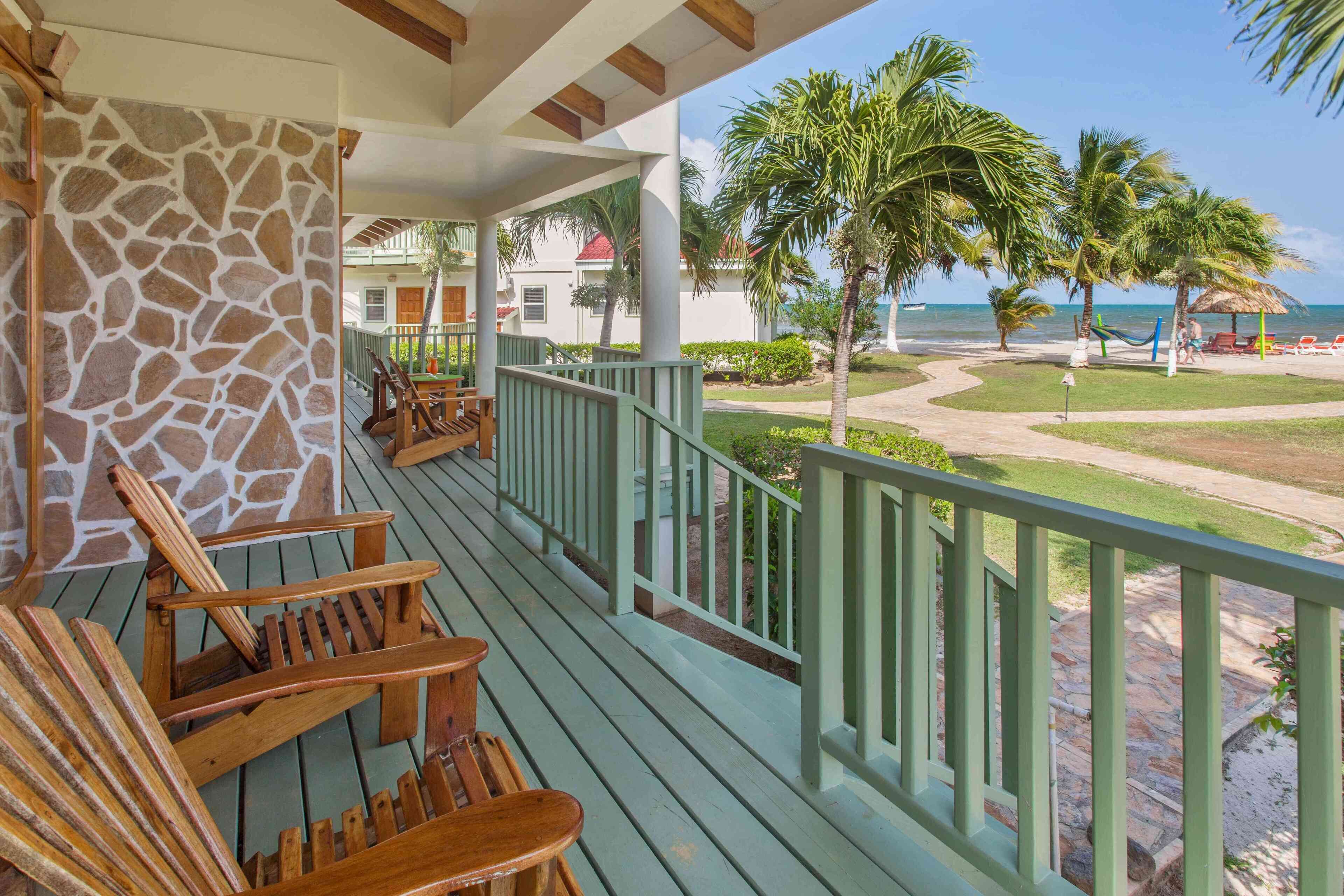 A wooden deck with chairs, a stone wall, and a view of a beach with palm trees and a thatched structure.