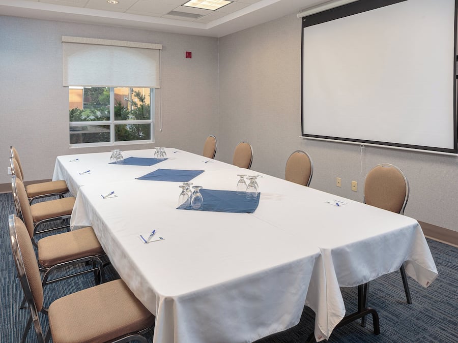 Un salón de conferencias con una larga mesa, sillas, una pantalla de proyector y un ventanal con vistas a zonas verdes.