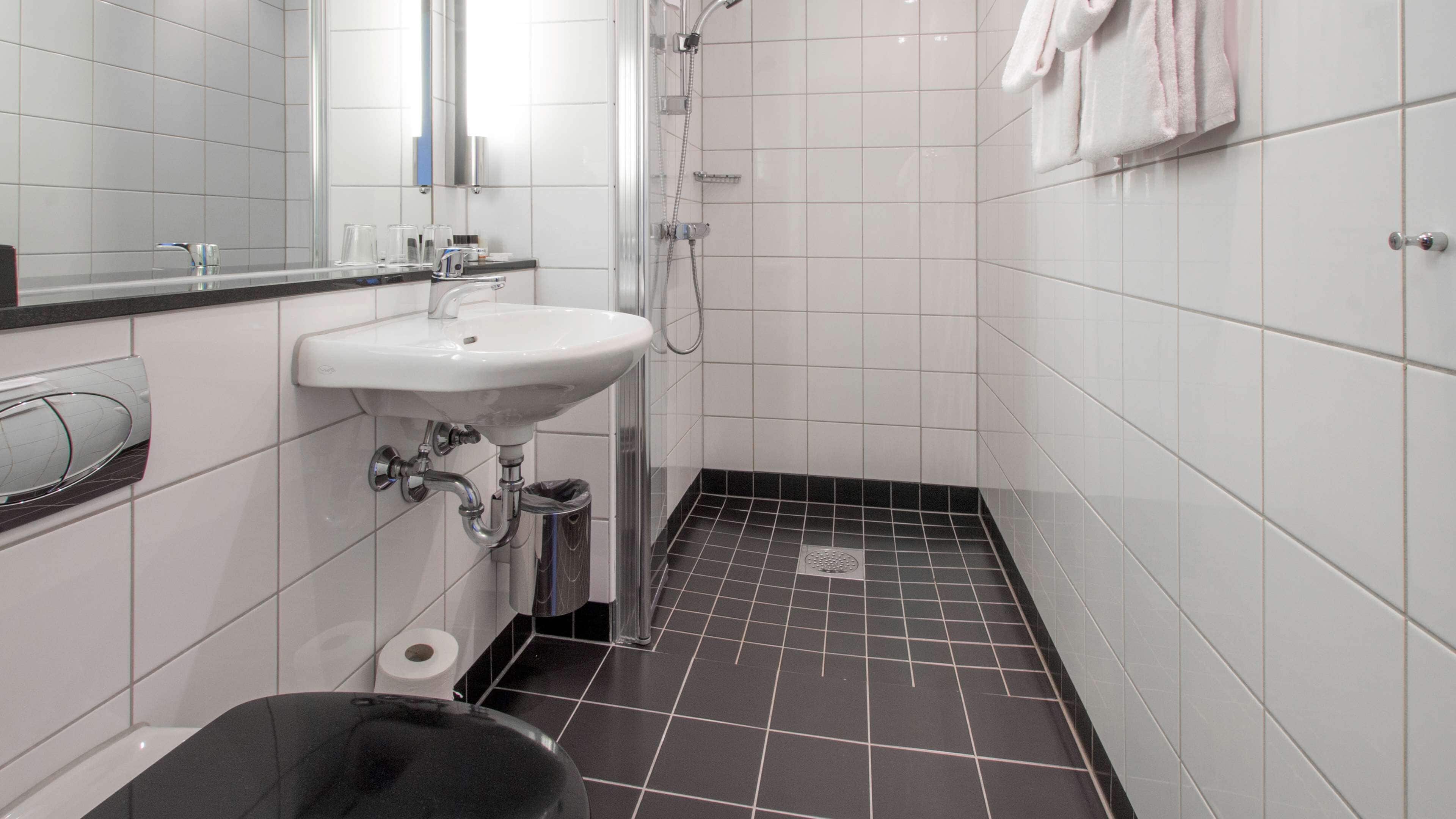 A small, modern bathroom with a white sink, a mirror, a shower, and a black trash bin.