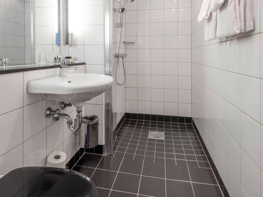 A small, modern bathroom with a white sink, a mirror, a shower, and a black trash bin.