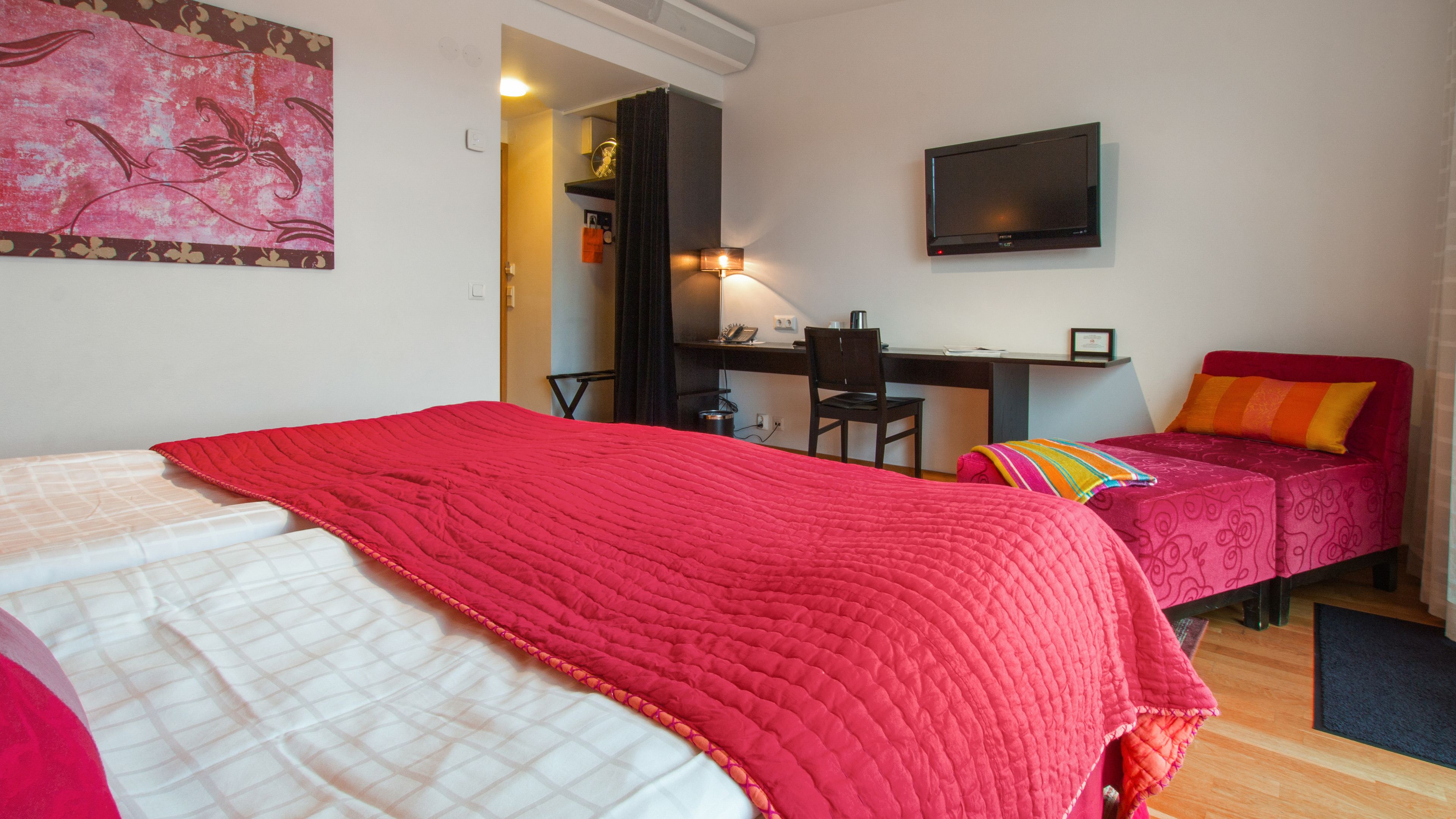 Een kamer met twee bedden, waarvan één met een rode sprei en de ander met een witte. Er is een bureau met een stoel, een aan de muur bevestigde televisie en een schilderij.