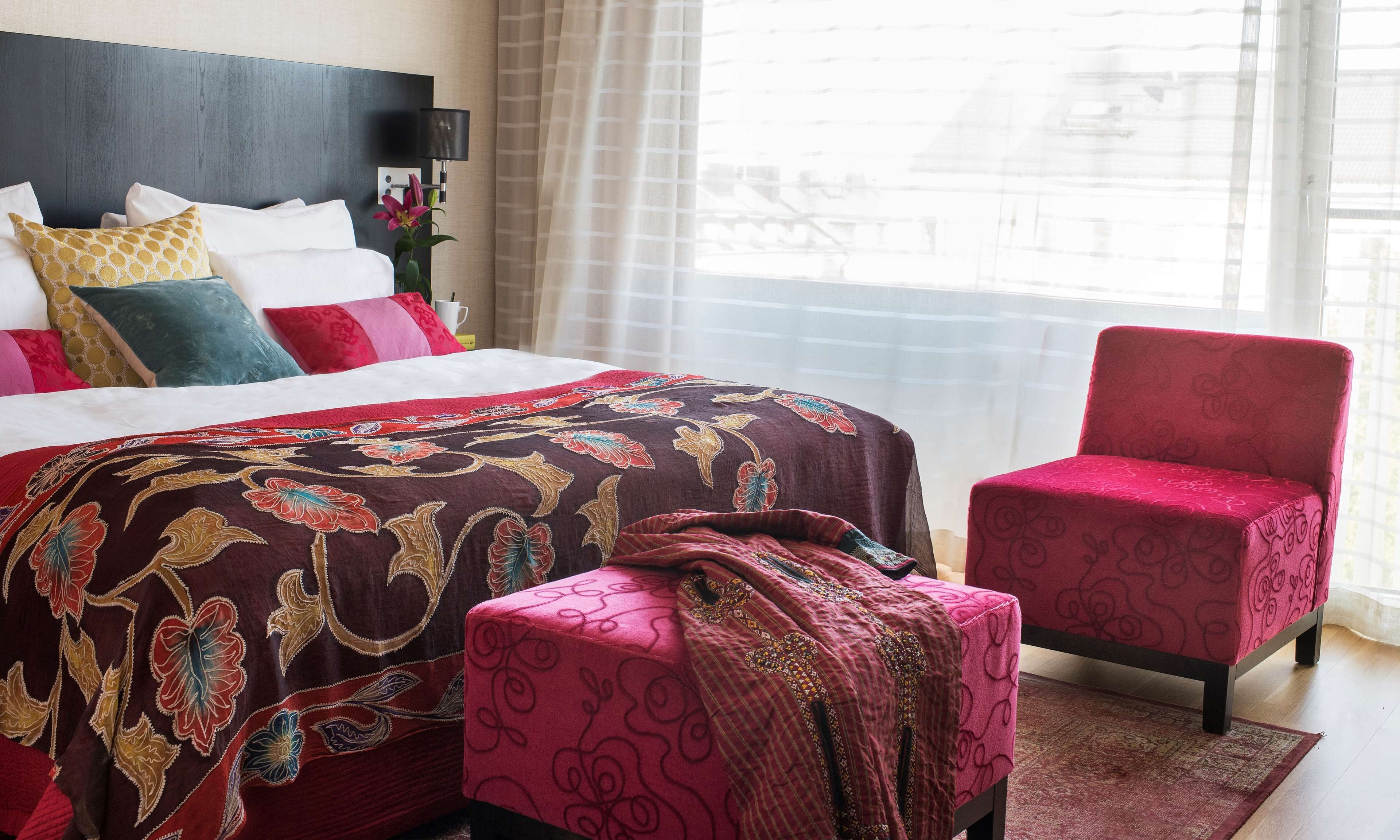 Een hotelkamer met een groot bed, een rode fauteuil, een klein voetenbankje en een nachtkastje met een lamp en een plant.