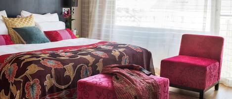 A hotel room with a large bed, a red armchair, a small ottoman, and a nightstand with a lamp and a plant.