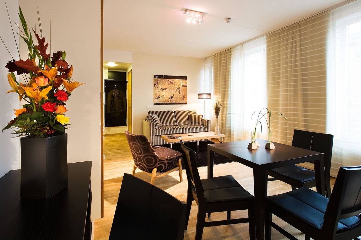 A modern living room with a striped sofa, a dining table with chairs, a vase of flowers, and a painting on the wall.