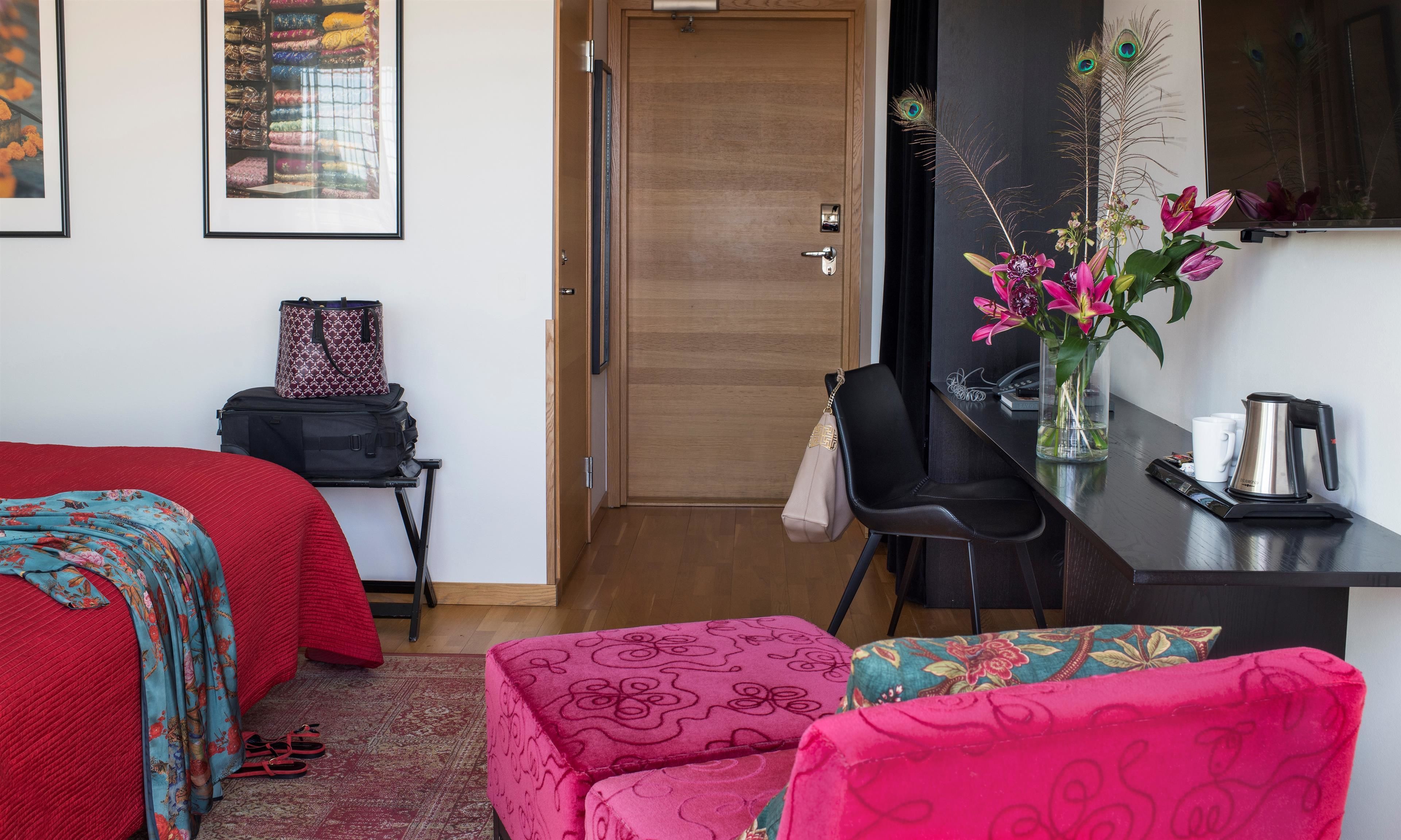 Habitación de hotel con una cama, un escritorio con una silla, una cafetera y un jarrón con flores.