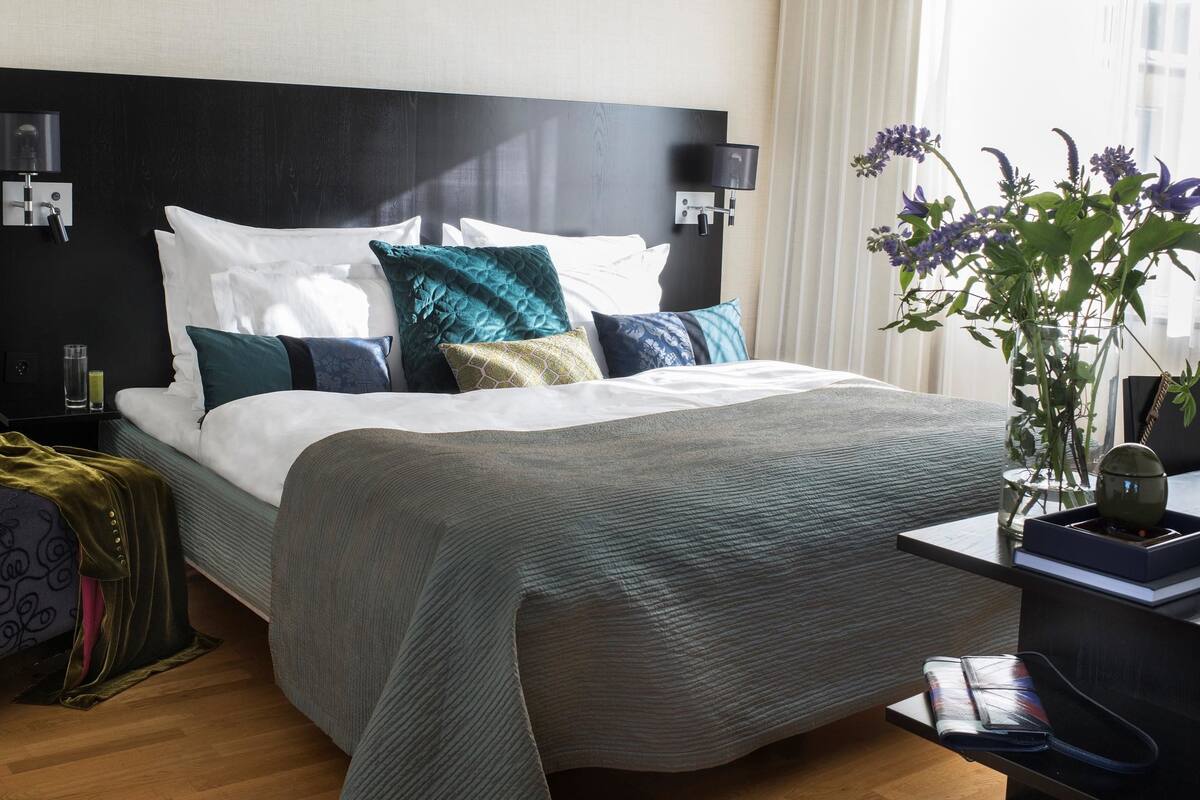 A modern hotel room with a large bed, a bedside table with a lamp, a chair, and a vase with flowers.