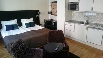 A hotel room with a bed, a desk, a chair, a remote control, and a kitchenette.