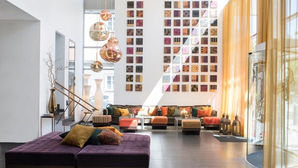 Amplia sala de estar con un amplio sofá, una mesita de centro y un jarrón decorativo. Se observan varias lámparas colgantes esféricas y una pared decorada con obras de arte estampadas.