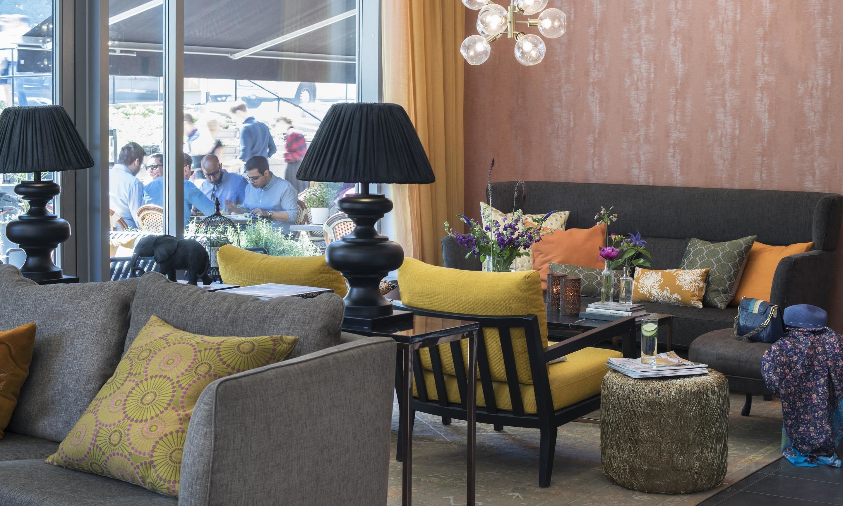 Un espacio lounge moderno con sofá, sillones y una mesa de centro. Hay lámparas decorativas, un jarrón con flores y una mesita con una bebida.