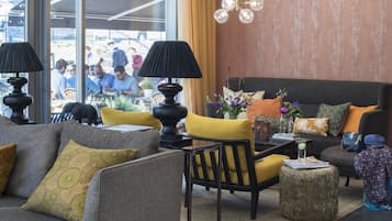 Un espacio lounge moderno con sofá, sillones y una mesa de centro. Hay lámparas decorativas, un jarrón con flores y una mesita con una bebida.