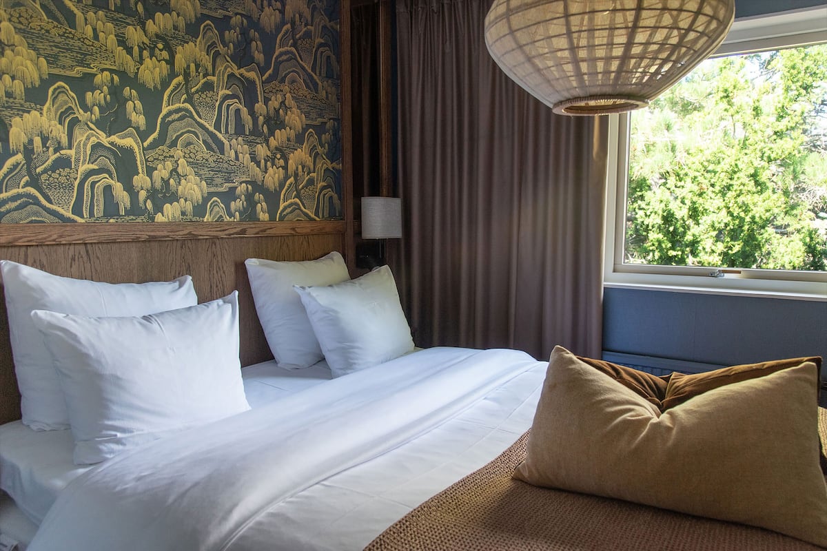 Habitación de hotel con cama, almohadas, una lámpara y vistas a zonas verdes por la ventana.