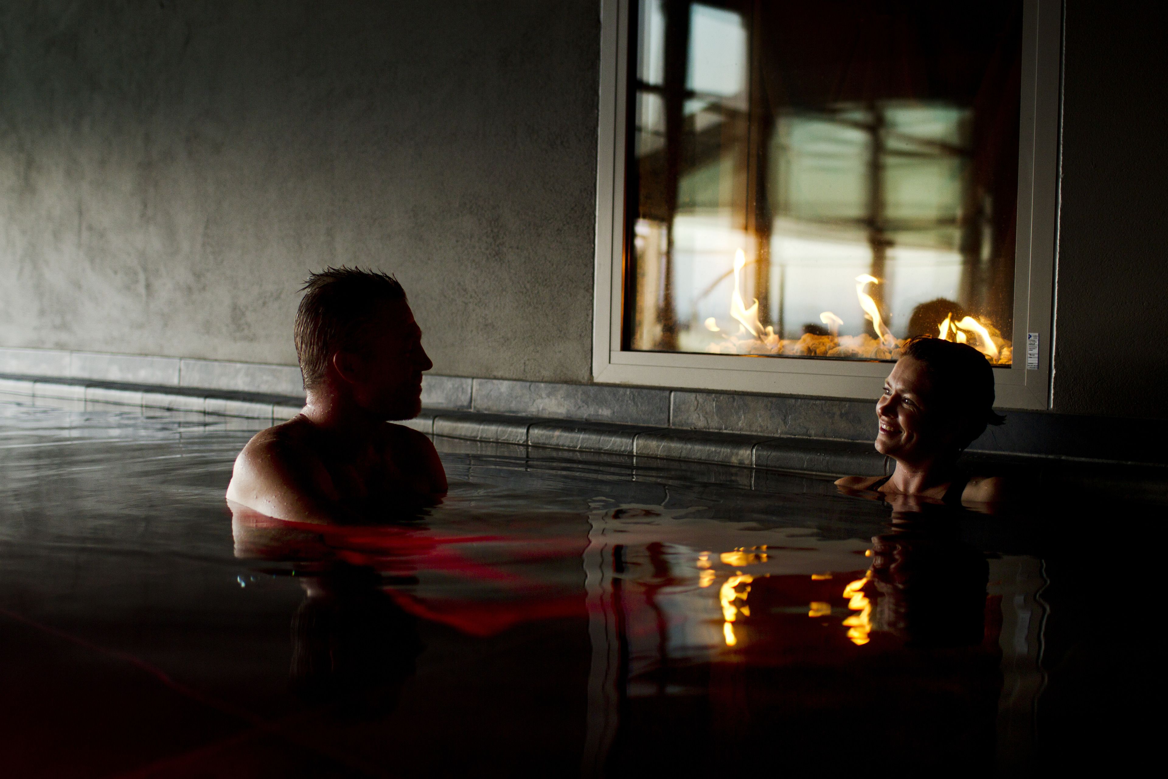 Dos personas en una piscina interior tenuemente iluminada con un amplio ventanal que refleja un fuego.