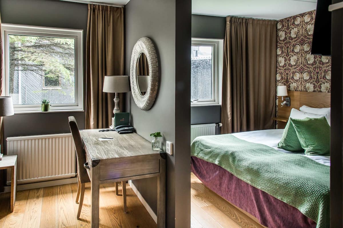 Una habitación con una mesa de madera, una silla, una lámpara, un espejo y una cama con ropa de cama verde.