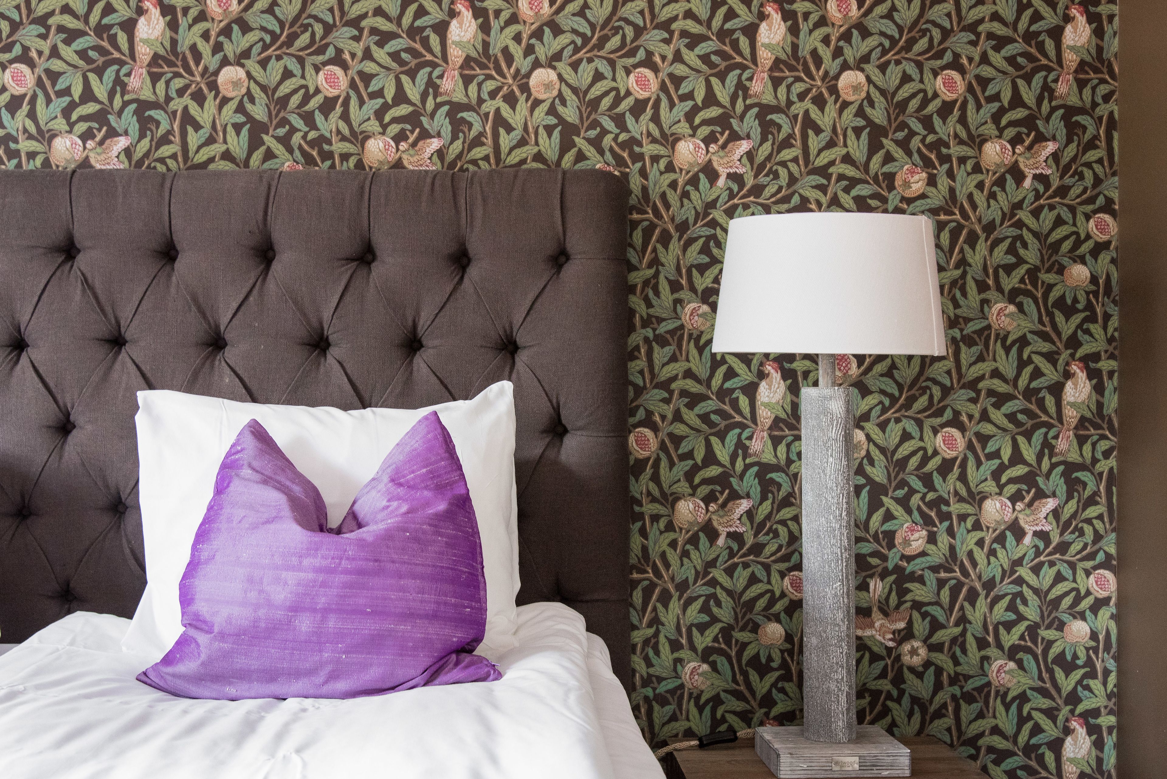 A bed with a tufted headboard, a white pillow, and a purple accent pillow. A floor lamp with a white shade is beside the bed. The wall has a floral wallpaper pattern.
