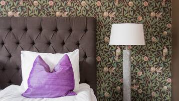 A bed with a tufted headboard, a white pillow, and a purple accent pillow. A floor lamp with a white shade is beside the bed. The wall has a floral wallpaper pattern.