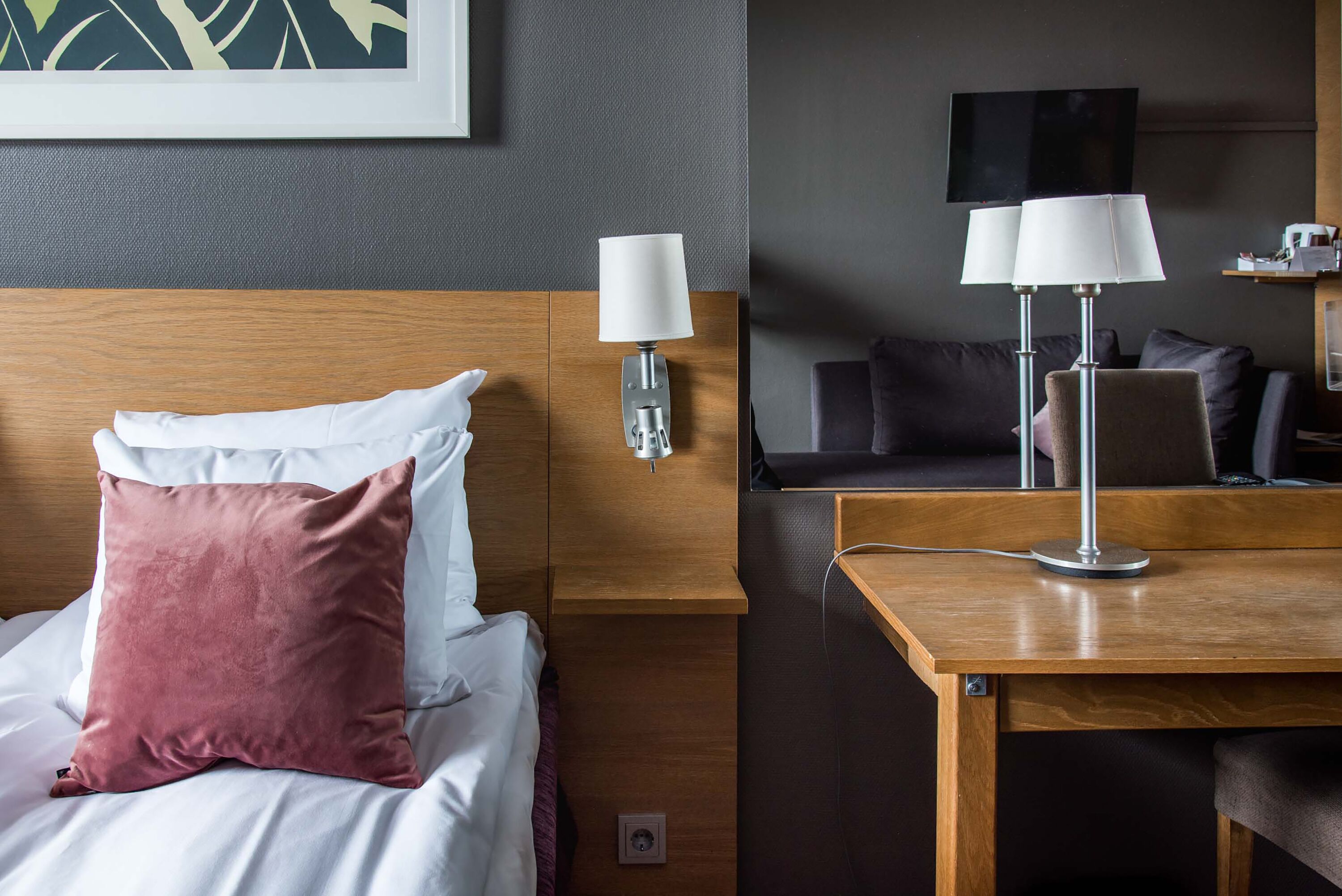 A hotel room with a bed, a nightstand with a lamp, a wooden desk, and a couch.