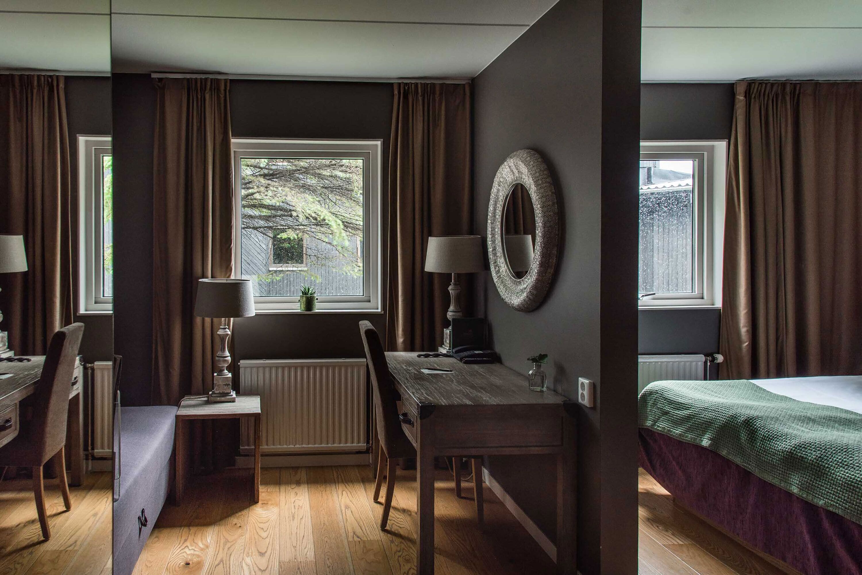 Una habitación con piso de madera, una mesa de comedor con sillas, una cama con manta verde, una ventana con cortinas y un espejo fijado a la pared.