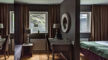 Una habitación con piso de madera, una mesa de comedor con sillas, una cama con manta verde, una ventana con cortinas y un espejo fijado a la pared.