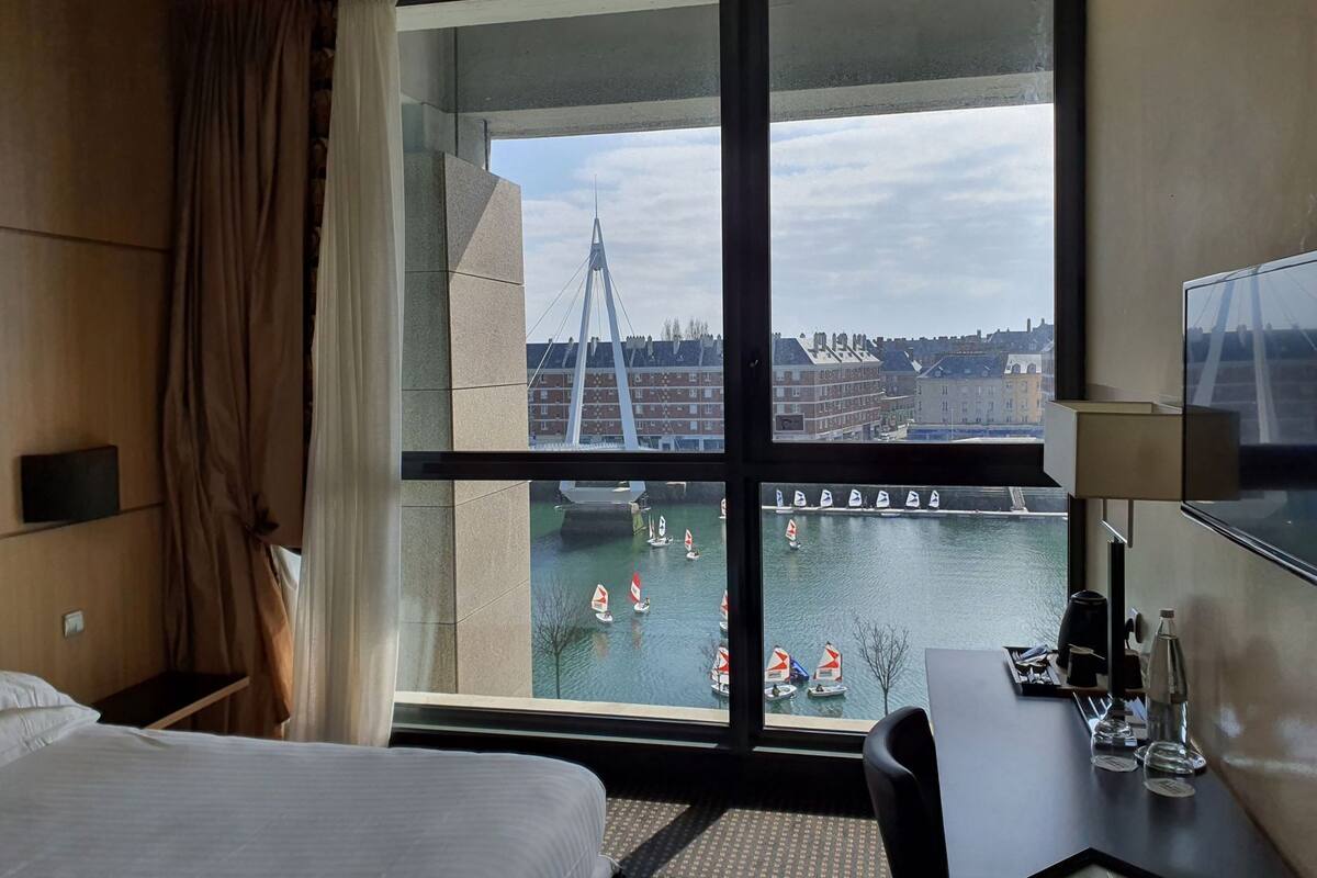 A hotel room with a large window, a bed, a desk, and a TV.