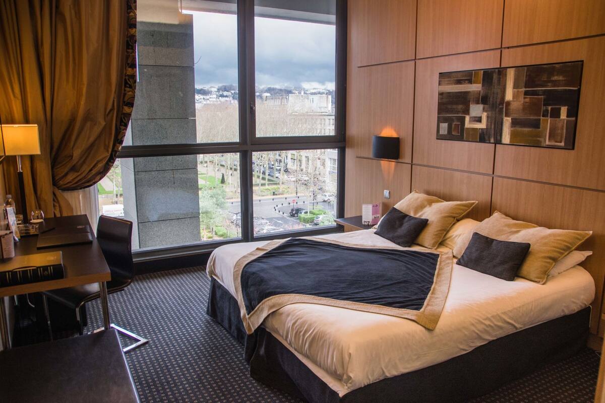 A hotel room with a large bed, a desk with a laptop, a lamp, and a view of the city through a large window.