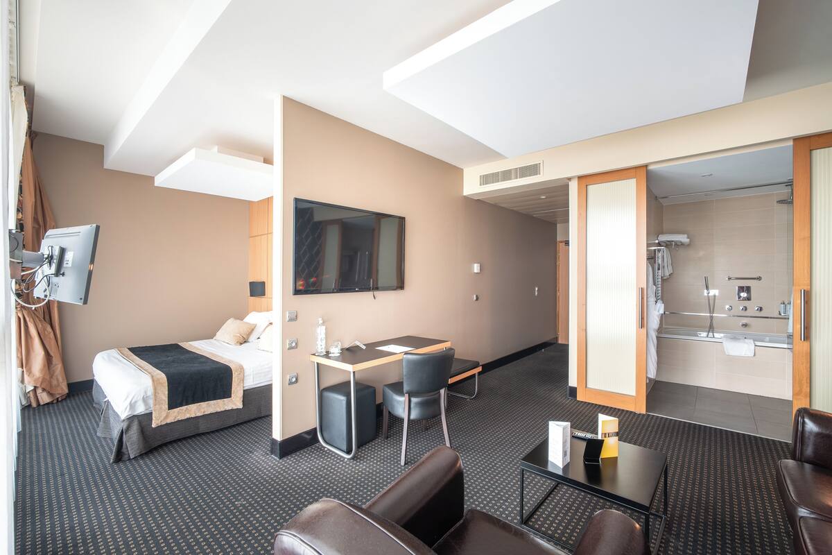 A hotel room with a bed, desk, two chairs, a TV, and a bathroom visible through an open door.