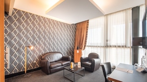 Una habitación de hotel moderna con un papel tapiz de diseño geométrico, un sofá de cuero, una mesa de centro, un escritorio con una silla y un ventanal con cortinas.