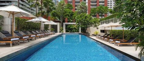 A swimming pool with lounge chairs and umbrellas, surrounded by greenery and tall buildings.