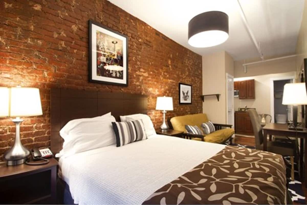 A hotel room with a brick wall, a large bed with white bedding, two bedside tables with lamps, a chair, and a desk.
