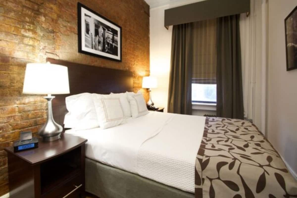 A hotel room with a brick wall, a bed with white bedding, a patterned throw, a nightstand with a lamp, and a framed picture above the bed.