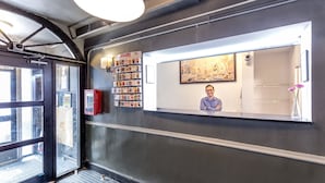 A reception area with a person seated behind a counter, a wall-mounted bookcase, and a framed cityscape painting.