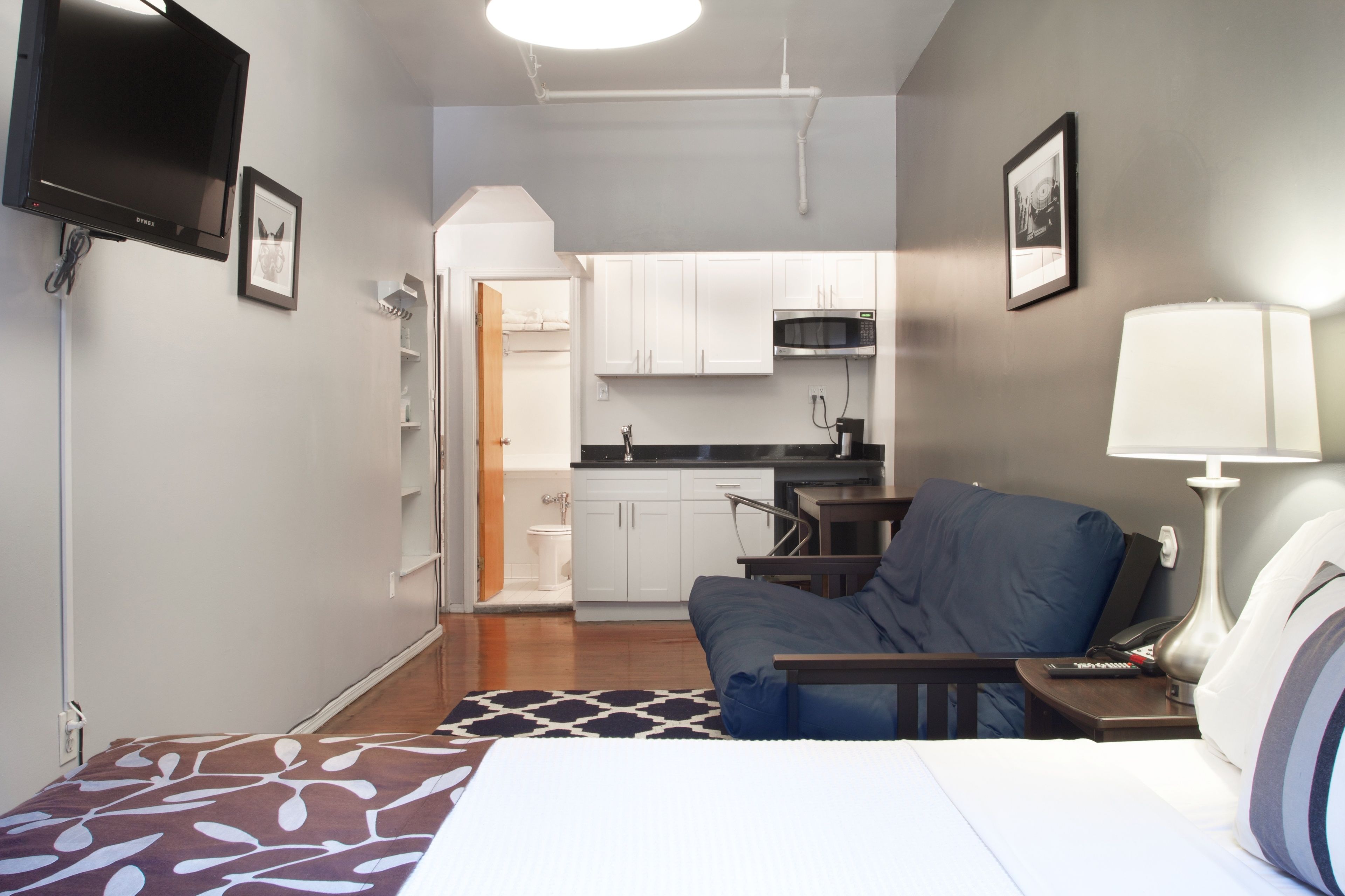 Ein kleines Hotelzimmer mit Bett, Schreibtisch, Stuhl, Lampe, Fernseher und einer Küchenzeile.