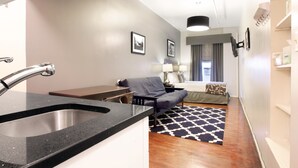 A compact hotel room with a kitchenette, a sofa, a bed, and a patterned rug.