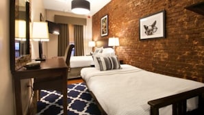 A hotel room with a brick wall, a bed with white bedding, a desk with a lamp, and a framed picture on the wall.