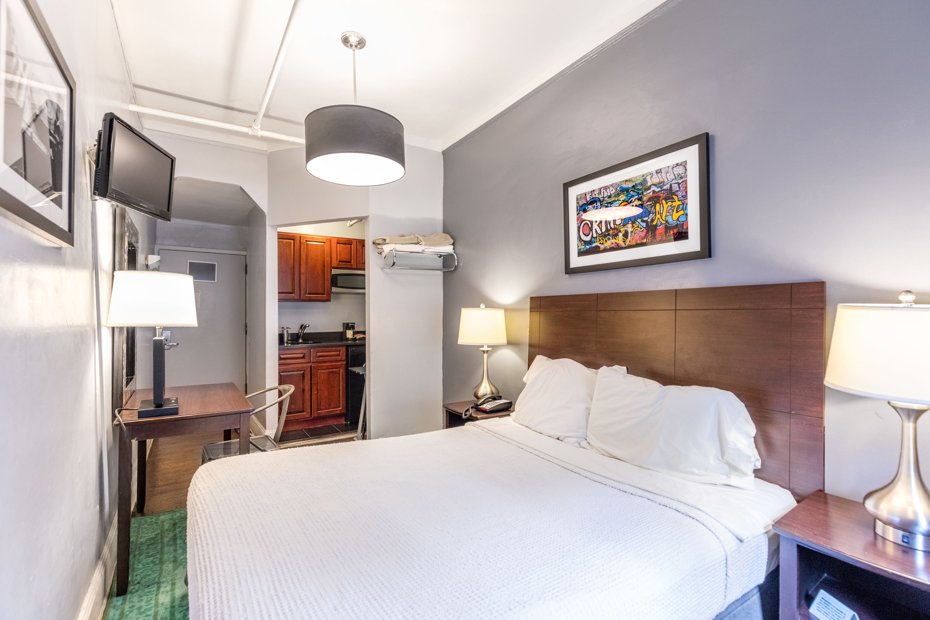 Une chambre d’hôtel avec un lit, des lampes de chevet, une télévision, un tableau encadré et une vue sur la cuisine.
