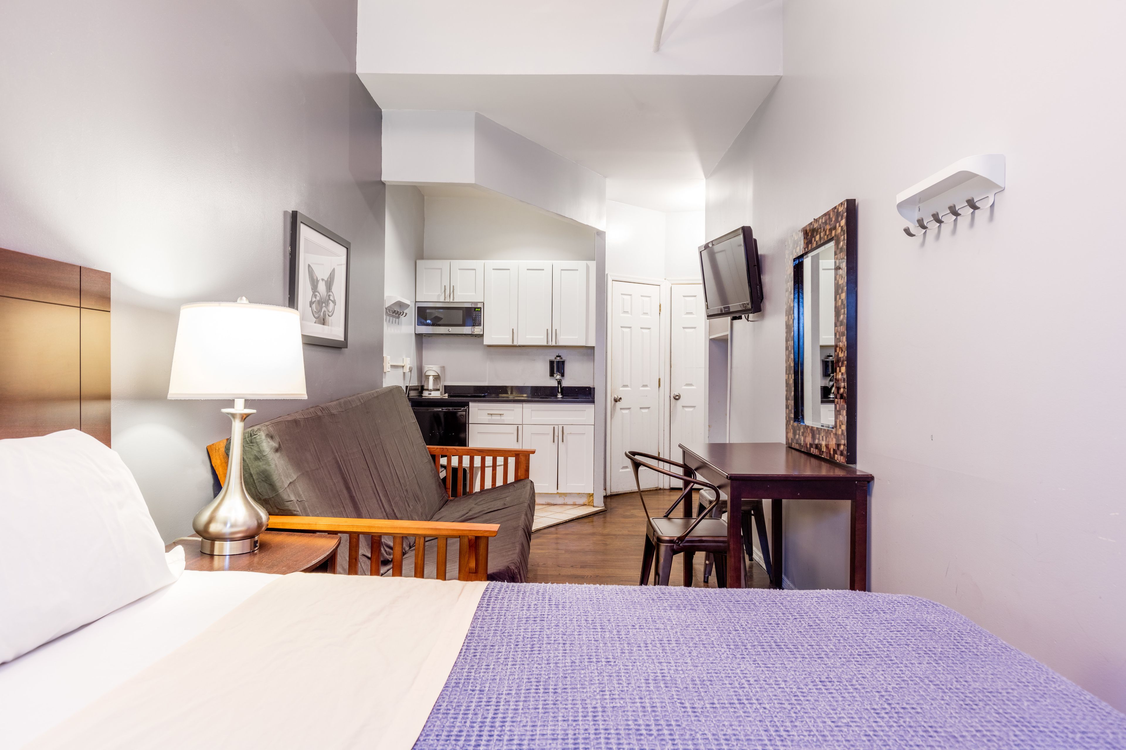 A hotel room with a bed, a desk, a chair, a lamp, a TV, a kitchen area, and a bathroom.