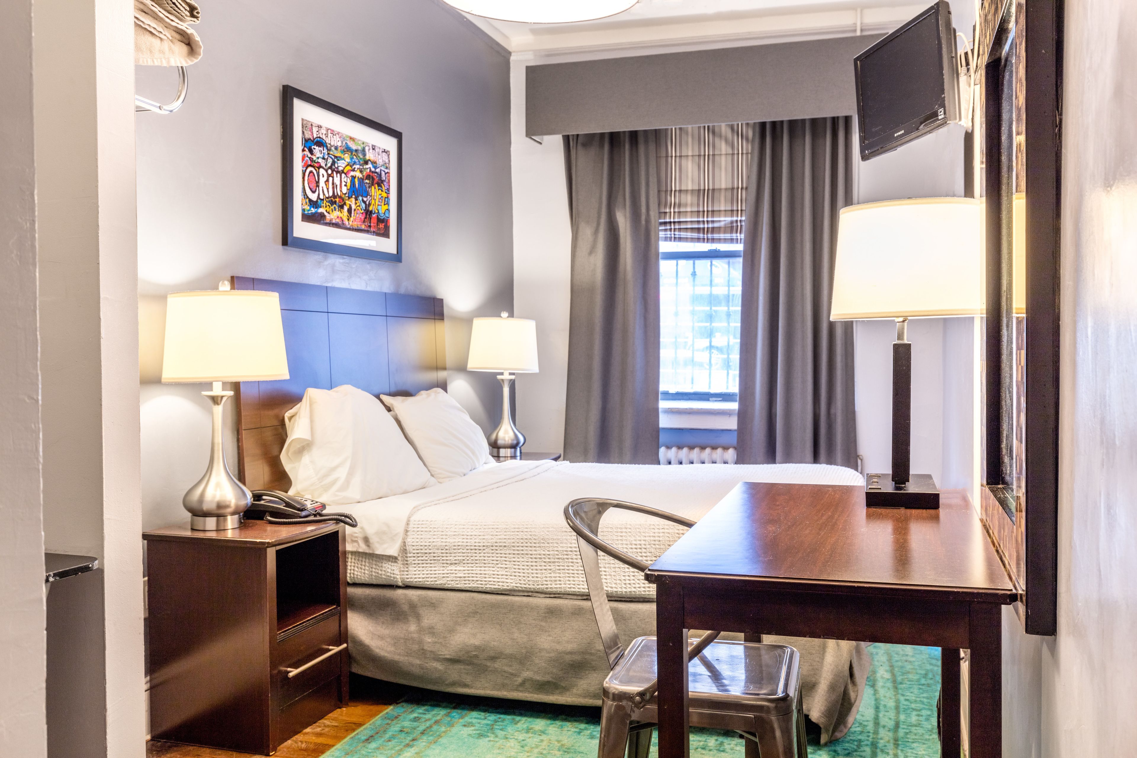 A hotel room with a bed, two bedside lamps, a desk, a chair, a framed picture on the wall, and a window with curtains.