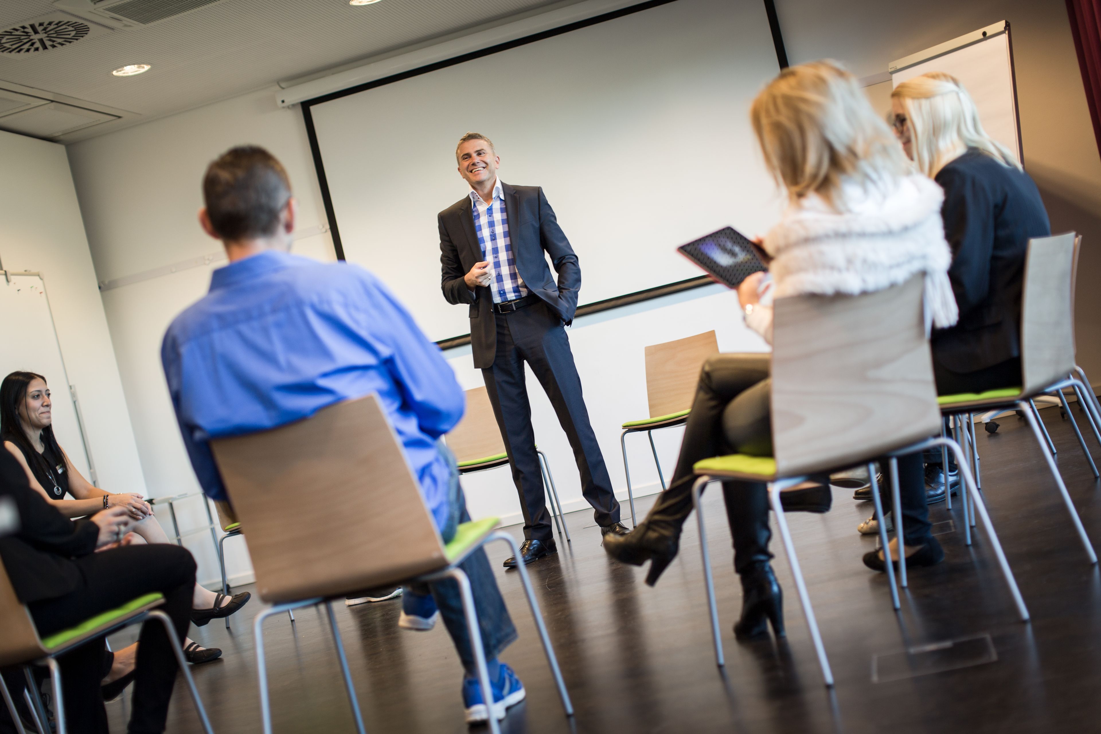 Sala de conferências com tela de apresentação, cadeiras dispostas em semicírculo e participantes voltados para o palestrante.