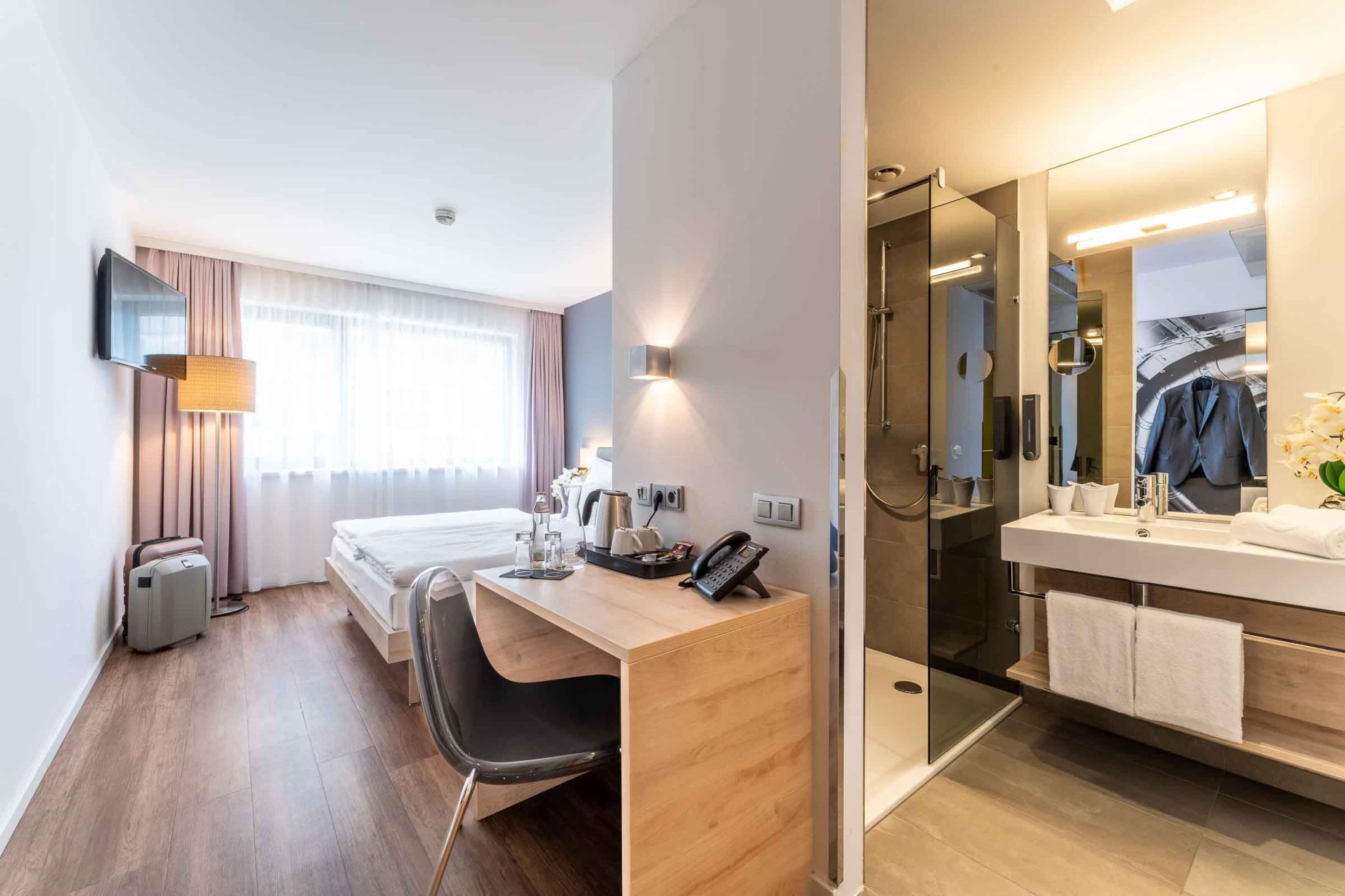 Habitación de hotel con cama, escritorio, silla, ducha y un espejo grande.
