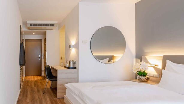 A hotel room with a bed, a desk, a chair, a round mirror, and a wall-mounted light fixture.