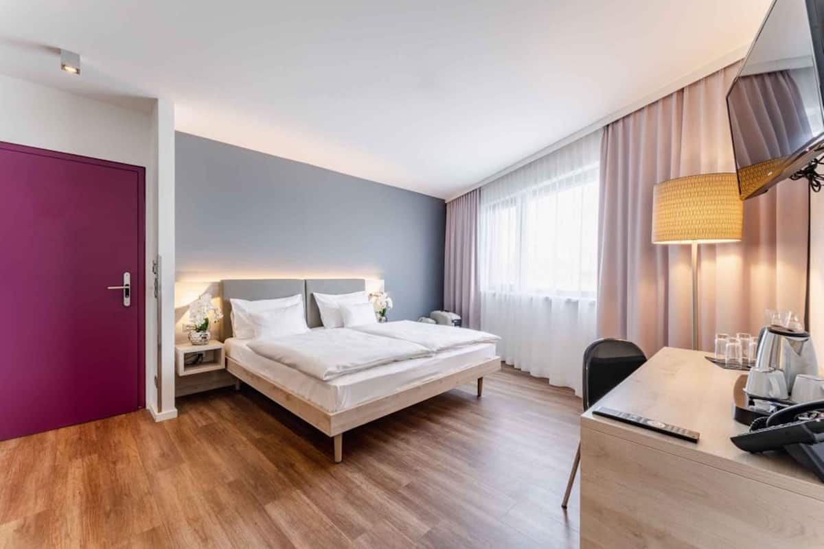 A modern hotel room with a wooden floor, a bed with white linens, a desk with a lamp, and a large window with curtains.