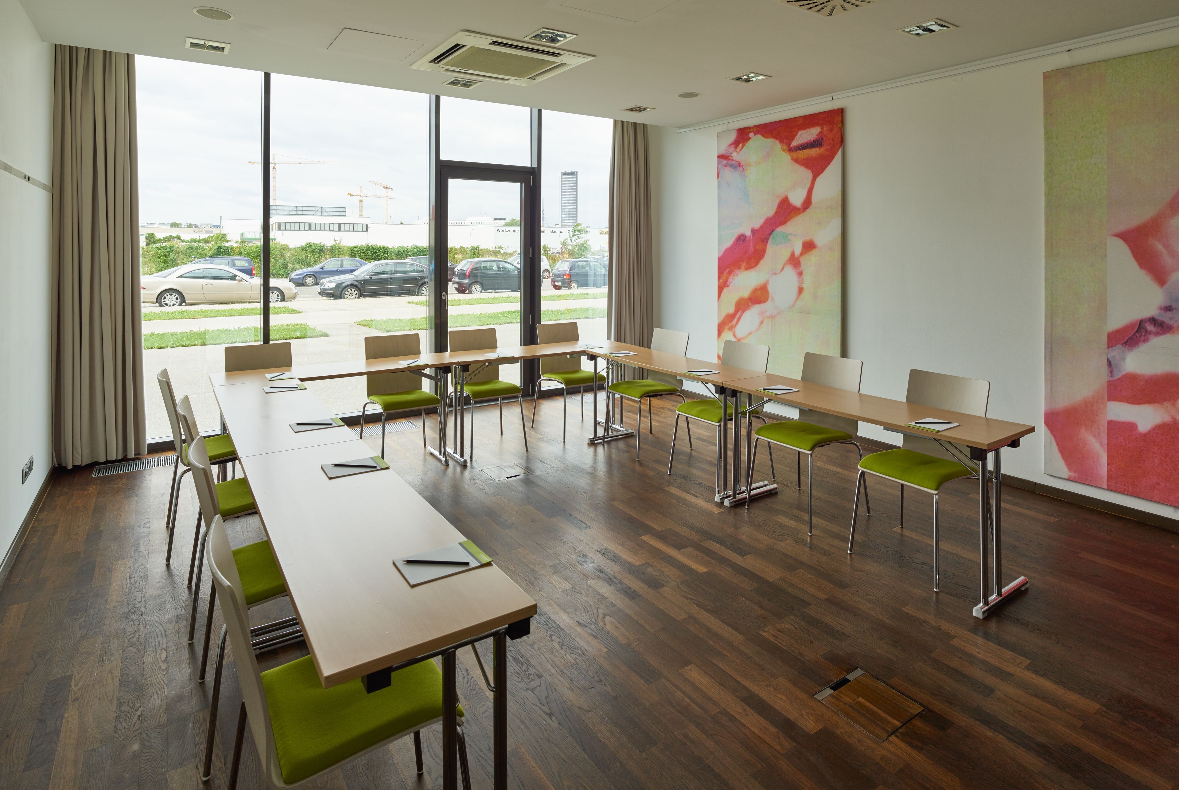A conference room with a long table and chairs, large windows with a view of parked cars and buildings, and a large abstract painting on the wall.