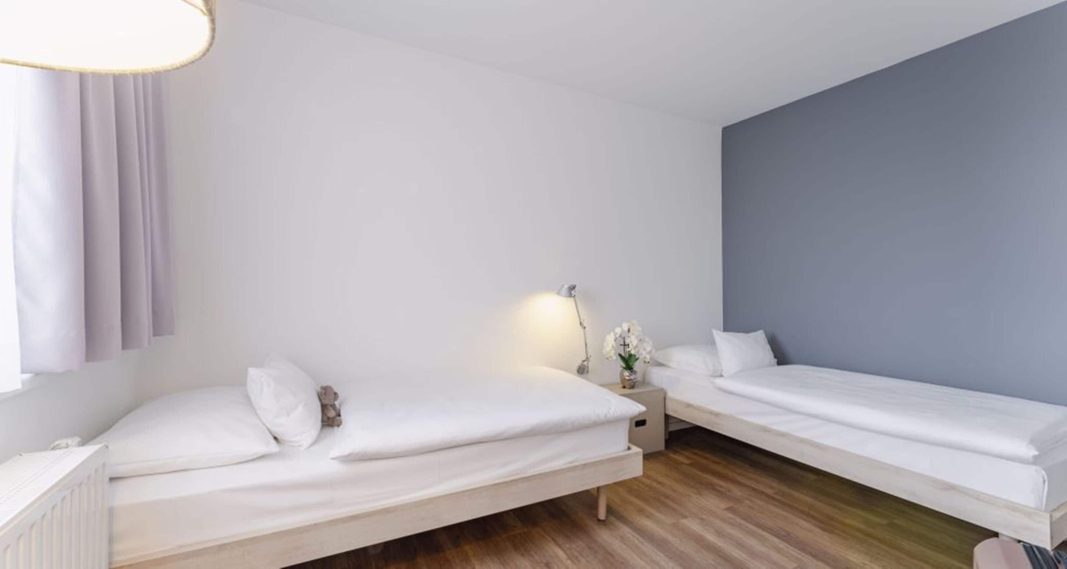 A modern hotel room with two single beds, a nightstand with a lamp, and a window with curtains.