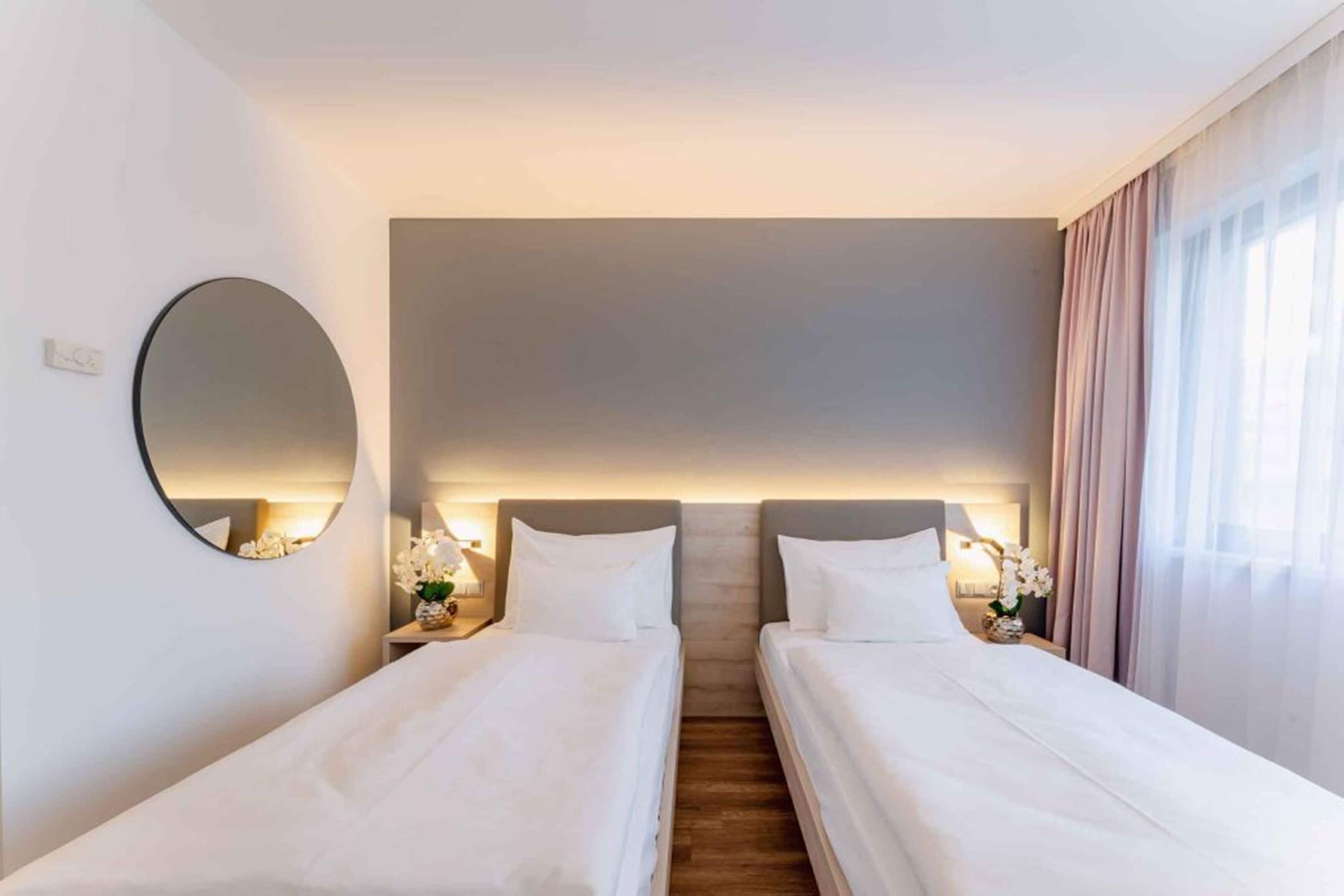 A hotel room with two single beds, a round mirror, bedside tables with lamps, and a view of the outdoors through a window with curtains.