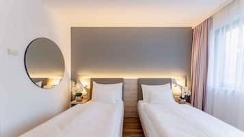 A hotel room with two single beds, a round mirror, bedside tables with lamps, and a view of the outdoors through a window with curtains.
