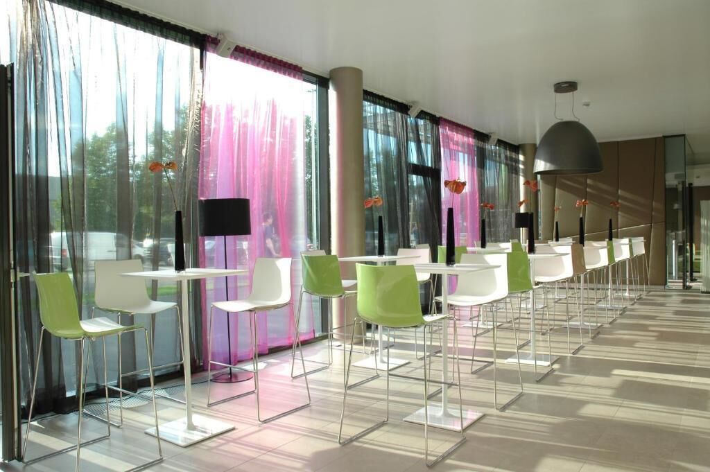 A modern indoor space with rows of white tables and green chairs, large windows with sheer curtains, and a minimalist design.