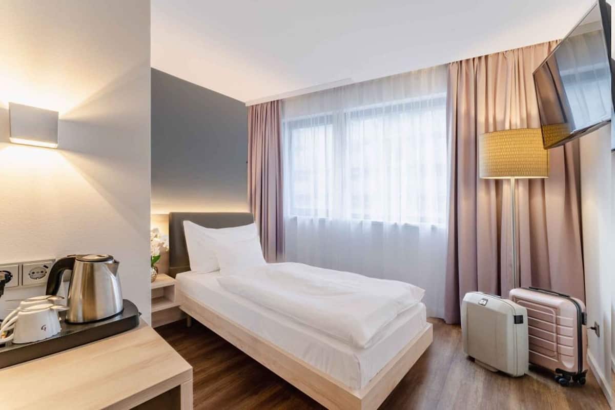 A hotel room with a bed, bedside table with a kettle, a lamp, curtains, and a TV.